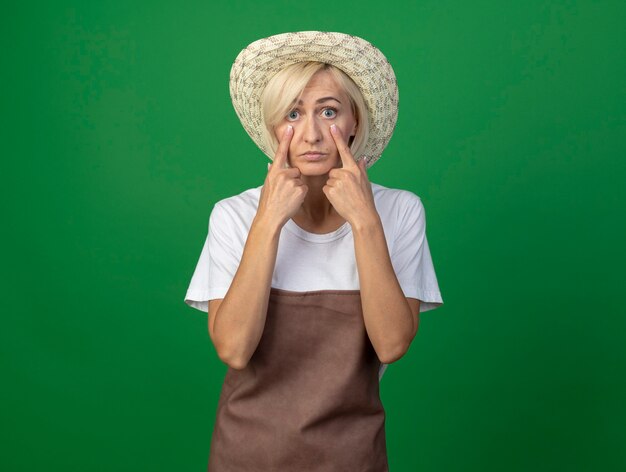 impressed middle-aged blonde gardener woman in uniform wearing hat  pulling down eye lids isolated on green wall with copy space