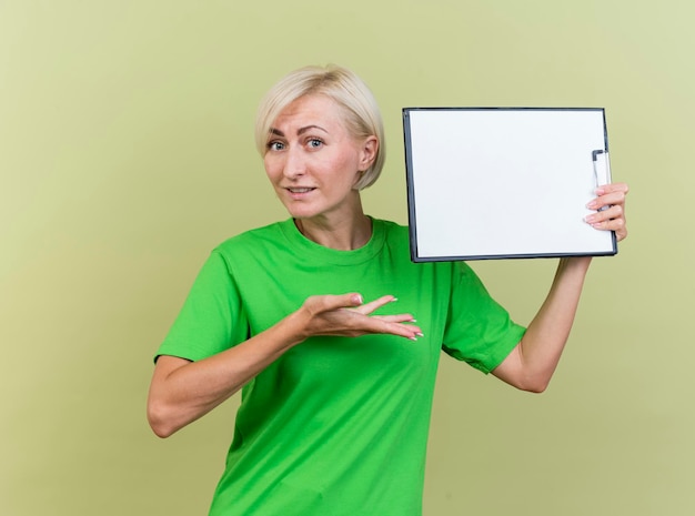 Впечатленная белокурая славянская женщина средних лет, держащая буфер обмена возле головы, указывая рукой на нее, смотрящую вперед, изолированную на оливково-зеленой стене