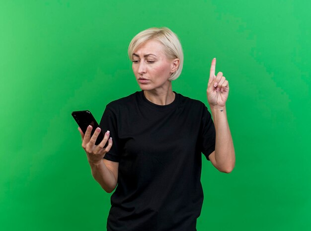 Impressed middle-aged blonde slavic woman holding and looking at mobile phone raising finger isolated on green wall with copy space