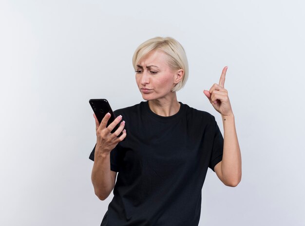 Впечатлила белокурая славянская женщина средних лет, держащая и смотрящая на мобильный телефон, поднимая палец на белом фоне с копией пространства