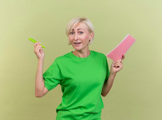 Impressed middle-aged blonde slavic woman holding pen and note pad looking at front isolated on olive green wall