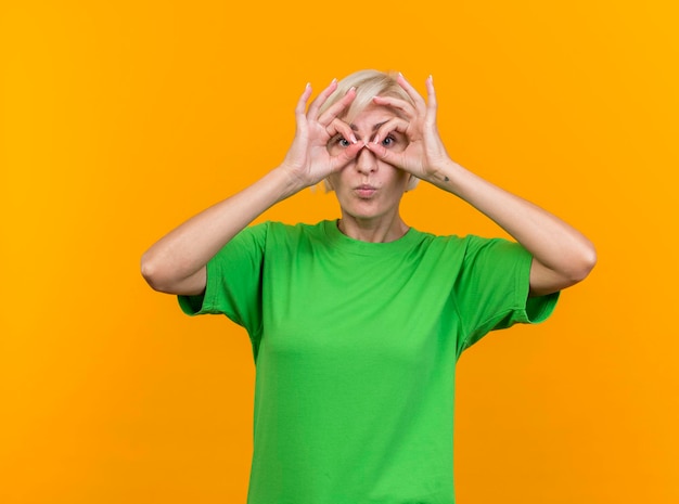 Foto gratuita donna slava bionda di mezza età colpita che guarda davanti facendo gesto di sguardo usando le mani come binocolo isolato sulla parete gialla