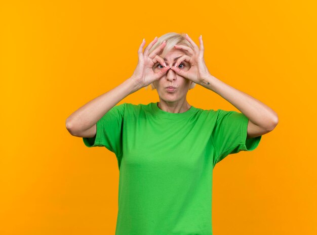 Impressed middle-aged blonde slavic woman looking at front doing look gesture using hands as binoculars isolated on yellow wall