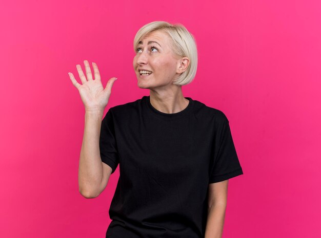 Impressed middle-aged blonde slavic woman looking at side doing hi gesture isolated on pink wall with copy space