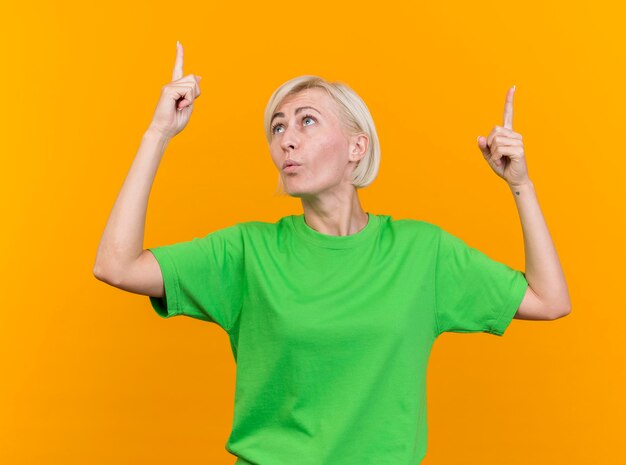 Впечатленная белокурая славянская женщина средних лет, указывая вверх, глядя на свой палец, изолированный на желтой стене
