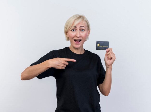 Impressed middle-aged blonde slavic woman showing credit card pointing at it looking at camera isolated on white background