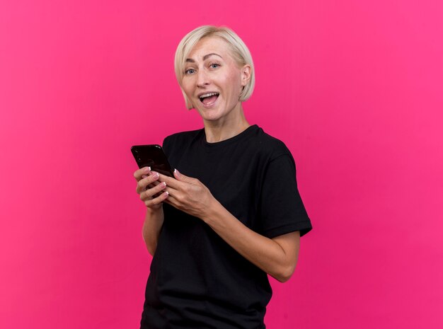 Impressed middle-aged blonde slavic woman standing in profile view holding mobile phone  isolated on crimson wall with copy space