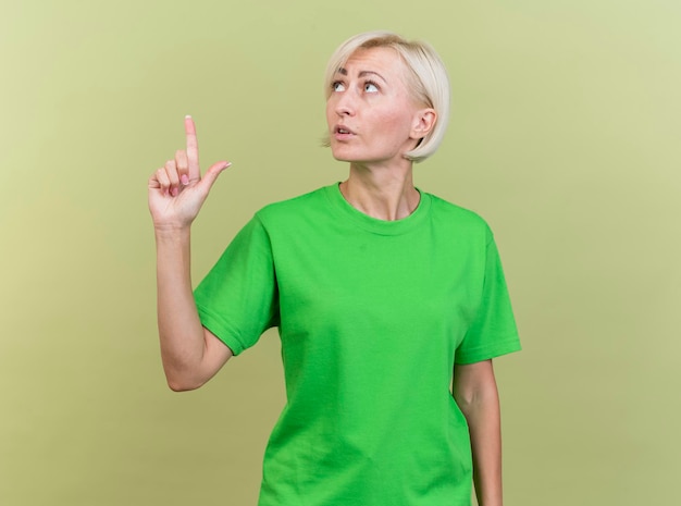 Impressed middle-aged blonde slavic woman turning head to side looking and pointing up isolated on olive green wall with copy space