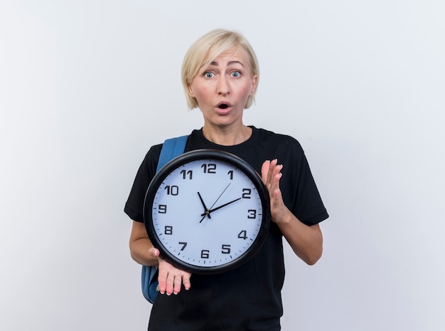 Impressed middle-aged blonde slavic woman wearing back pack holding clock looking at camera isolated on white background with copy space