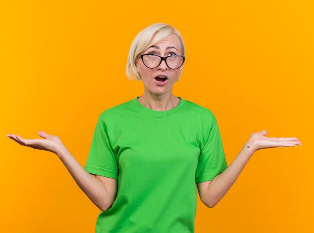 Impressed middle-aged blonde slavic woman wearing glasses showing empty hands looking up isolated on yellow wall