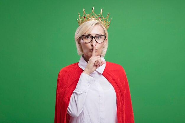Impressed middle-aged blonde superhero woman in red cape wearing glasses and crown doing silence gesture 