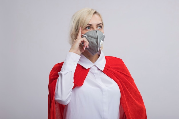 Impressed middle-aged blonde superhero woman in red cape wearing protective mask looking at side doing think gesture isolated on white wall with copy space