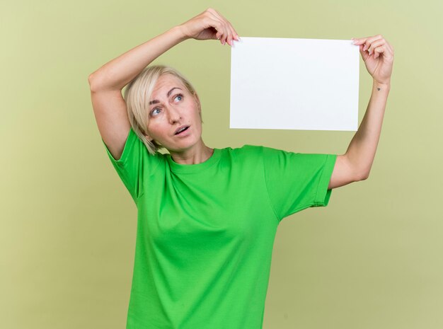 Бесплатное фото Впечатленная блондинка средних лет, держащая чистый лист бумаги возле головы, глядя на нее, изолированную на оливково-зеленой стене