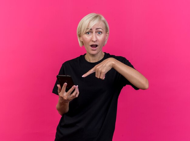 Impressed middle-aged blonde woman holding and pointing at mobile phone looking at front isolated on pink wall