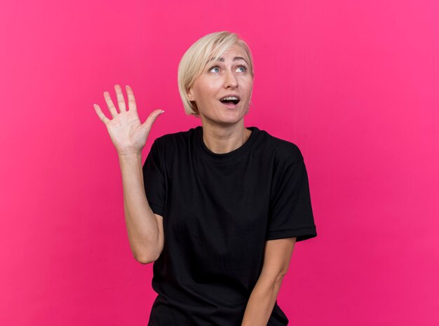 Impressed middle-aged blonde woman looking at side doing hi gesture isolated on pink wall