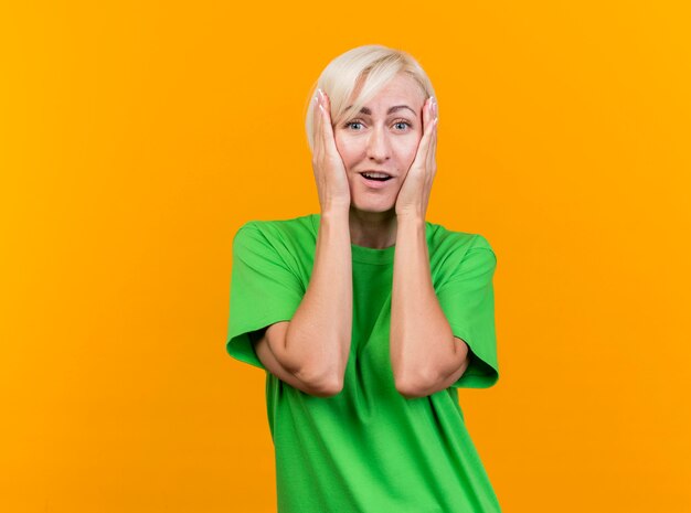 Impressed middle-aged blonde woman putting hands on face looking at front isolated on yellow wall