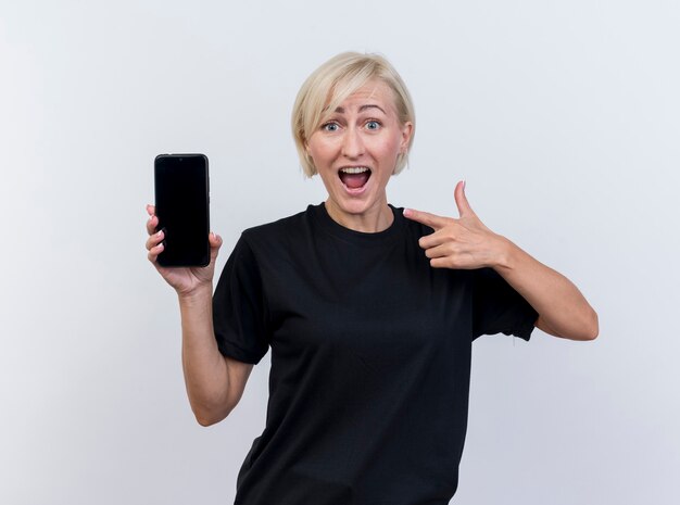 Впечатленная блондинка средних лет показывает мобильный телефон, указывая на него, глядя на переднюю часть, изолированную на белой стене