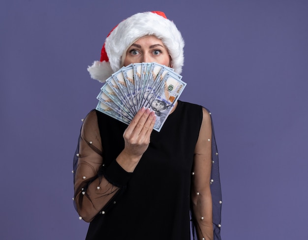 Impressed middle-aged blonde woman wearing christmas hat holding money looking at camera from behind it isolated on purple background