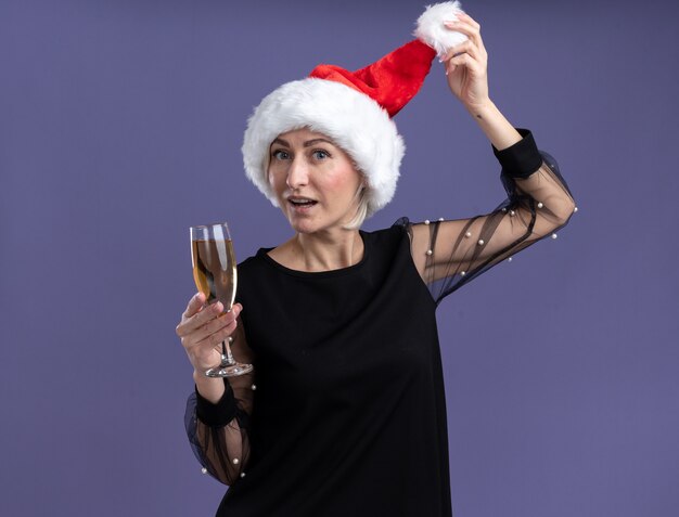 紫色の背景に分離されたシャンパンをつかむ帽子のガラスを保持しているカメラを見てクリスマス帽子をかぶっている印象的な中年のブロンドの女性