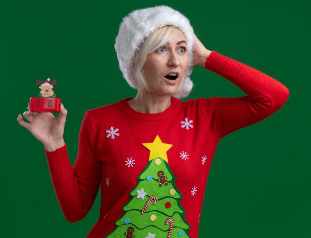 녹색 배경에 고립 된 머리에 손을 유지 측면에서 찾고 날짜와 크리스마스 순록 장난감을 들고 크리스마스 모자와 스웨터를 입고 감동 중년 금발의 여자