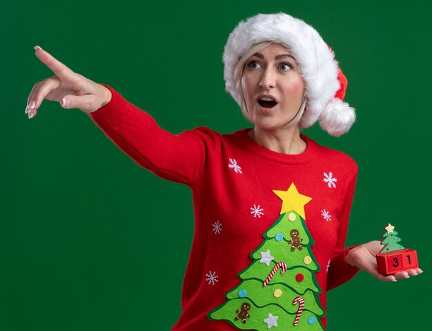 Impressed middle-aged blonde woman wearing christmas hat and sweater holding christmas tree toy with date looking and pointing at side isolated on green background