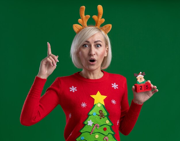 Impressed middle-aged blonde woman wearing christmas reindeer antlers headband and christmas sweater holding christmas reindeer toy with date looking at camera pointing up isolated on green background