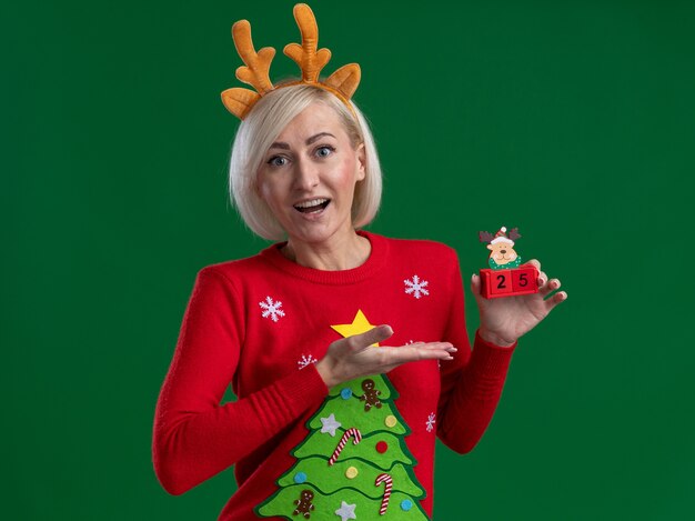 크리스마스 순록 뿔 머리띠와 크리스마스 스웨터를 입고 녹색 벽에 고립 된 날짜 찾고 크리스마스 순록 장난감에서 손으로 가리키는 감동 중년 금발의 여자