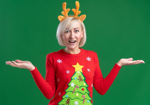 Impressed middle-aged blonde woman wearing christmas reindeer antlers headband and christmas sweater looking at camera showing empty hands isolated on green background