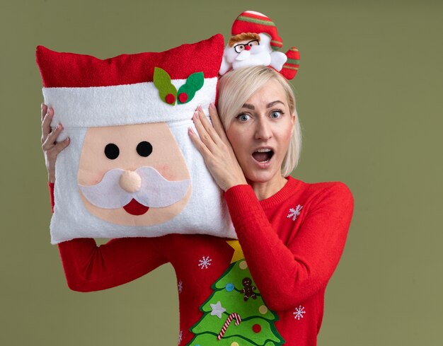 Впечатленная блондинка средних лет в головной повязке санта-клауса и рождественском свитере держит подушку санта-клауса, касаясь ею головы, глядя в камеру, изолированную на оливково-зеленом фоне