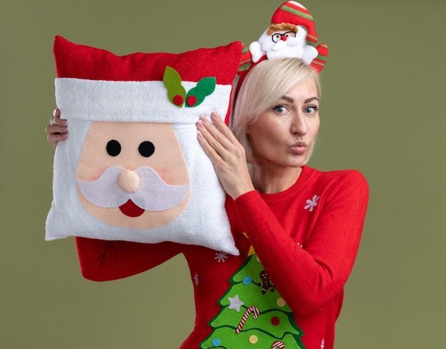 Free photo impressed middle-aged blonde woman wearing santa claus headband and christmas sweater holding santa claus pillow touching head with it looking  with pursed lips isolated on olive green wall