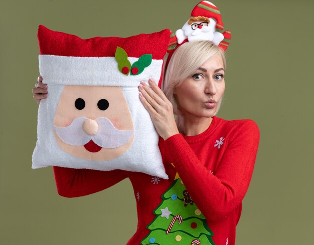 impressed middle-aged blonde woman wearing santa claus headband and christmas sweater holding santa claus pillow touching head with it looking  with pursed lips isolated on olive green wall