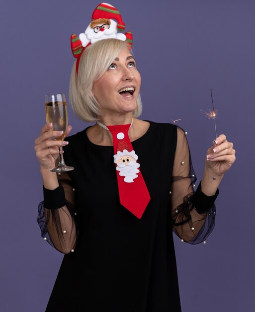 Impressed middle-aged blonde woman wearing santa claus headband and tie holding holiday sparkler and glass of champagne looking up isolated on purple background