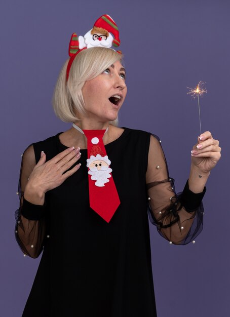 Impressed middle-aged blonde woman wearing santa claus headband and tie holding holiday sparkler keeping hand on chest looking up isolated on purple wall