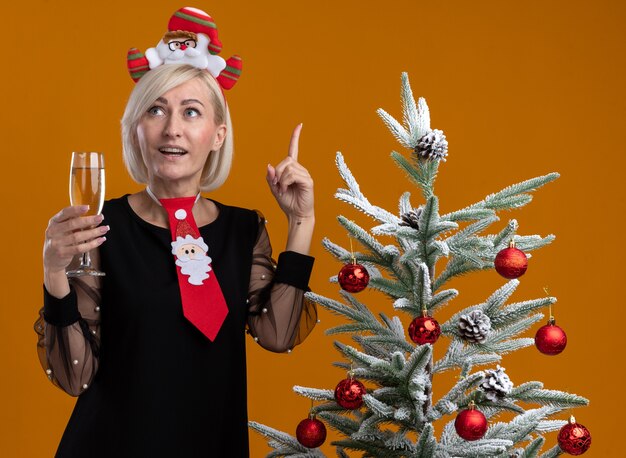 Impressed middle-aged blonde woman wearing santa claus headband and tie standing near decorated christmas tree holding glass of champagne looking and pointing up isolated on orange background