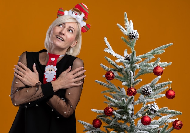 Impressed middle-aged blonde woman wearing santa claus headband and tie standing near decorated christmas tree keeping hands crossed on arms looking up isolated on orange wall