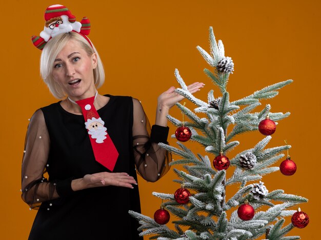 Impressed middle-aged blonde woman wearing santa claus headband and tie standing near decorated christmas tree pointing at it with hands looking at camera isolated on orange background