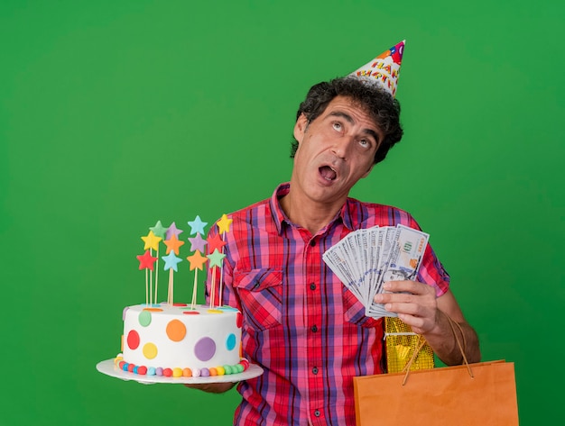 Impressed middle-aged caucasian party man wearing birthday cap holding birthday cake paper bag gift pack and money looking up isolated on green background