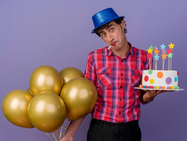 Impressionato uomo caucasico di mezza età che indossa il cappello del partito che tiene palloncini e torta di compleanno con ventilatore di partito in bocca che guarda l'obbiettivo isolato su priorità bassa viola