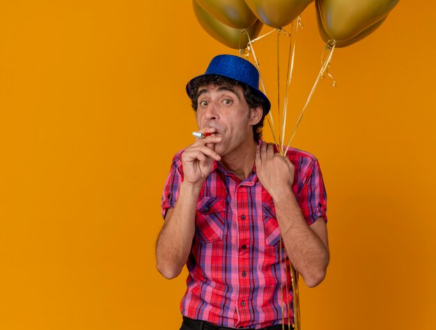 Impressed middle-aged caucasian party man wearing party hat holding balloons looking at camera holding party blower in mouth isolated on orange background with copy space