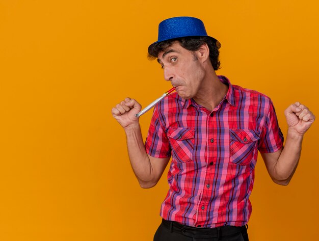 Impressed middle-aged caucasian party man wearing party hat looking at camera keeping fists in air blowing party blower isolated on orange background with copy space