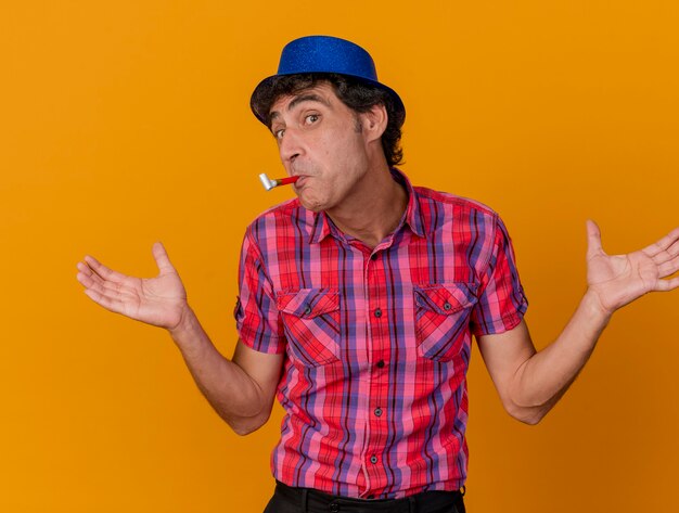 Impressed middle-aged caucasian party man wearing party hat looking at camera showing empty hands with party blower in mouth isolated on orange background