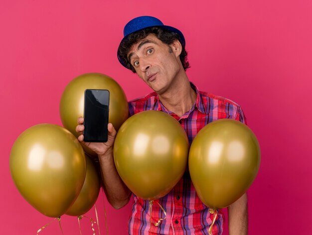 진홍색 배경에 고립 된 카메라를보고 휴대 전화를 보여주는 풍선 뒤에 서있는 파티 모자를 쓰고 감동 중년 백인 파티 남자