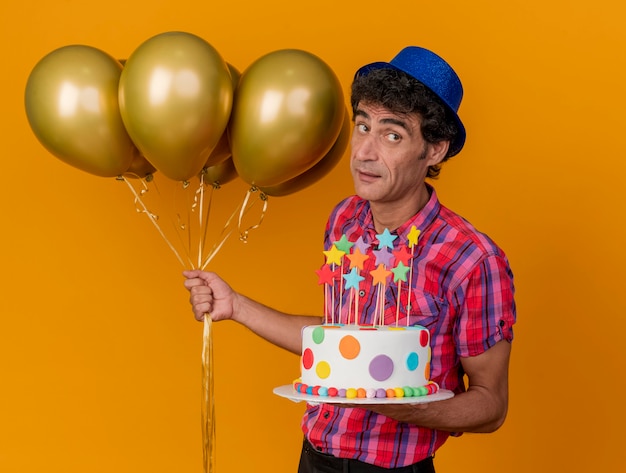 Free photo impressed middle-aged caucasian party man wearing party hat standing in profile view holding balloons and birthday cake looking at side isolated on orange background