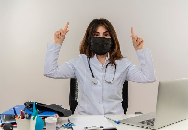 의료 도구 클립 보드와 노트북이 격리 가리키는 책상에 앉아 의료 마스크를 쓰고 감동 된 중년 여성 의사