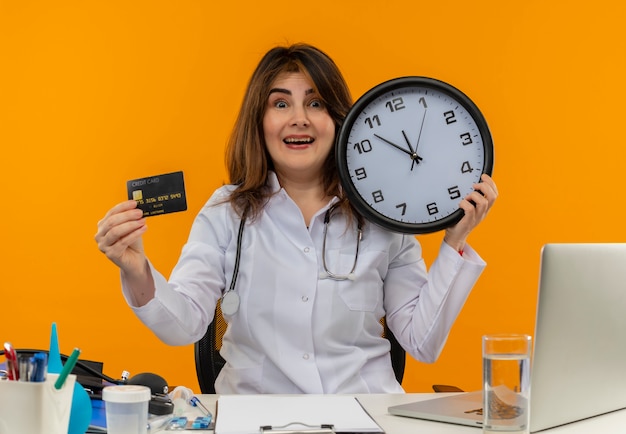 Impressed middle-aged female doctor wearing medical robe and stethoscope sitting at desk with medical tools clipboard and laptop holding clock and credit card isolated