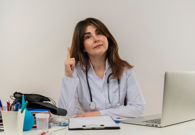 Impressed middle-aged female doctor wearing medical robe and stethoscope sitting at desk with medical tools clipboard and laptop raising finger looking isolated