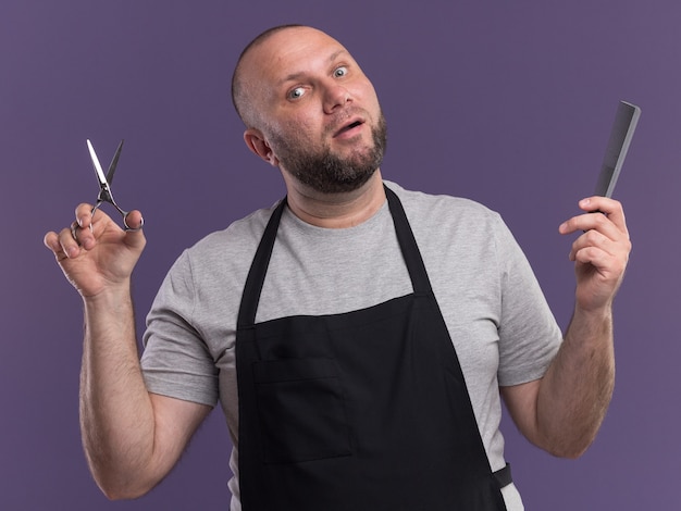 Impressed middle-aged male barber in uniform holding scissors with comb isolated on purple wall