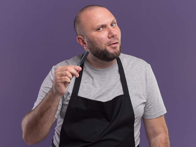 Impressed middle-aged male barber in uniform holding straight razor isolated on purple wall