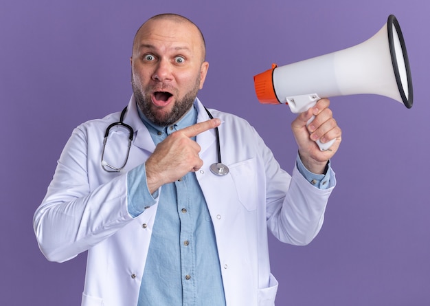 Impressed middle-aged male doctor wearing medical robe and stethoscope holding and pointing at speaker 