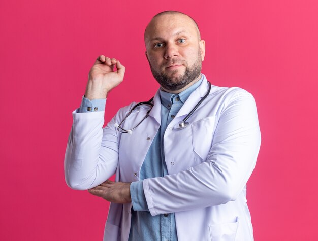 Впечатленный мужчина-врач средних лет в медицинском халате и стетоскопе, держащий руку в воздухе, изолированную на розовой стене
